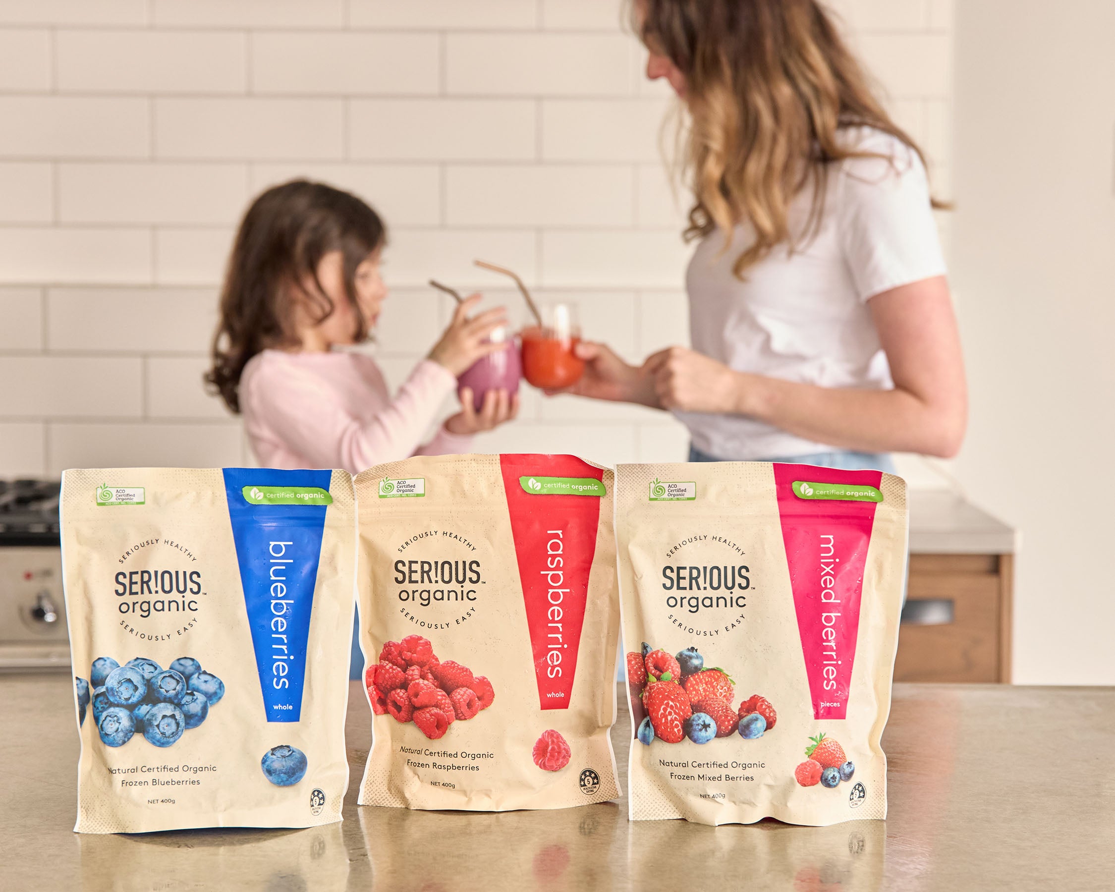Mother and daughter cheers smoothies behind Serious Organic frozen fruit range packs on table in kitchen.